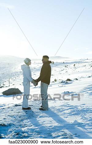 Stock Image - Male and female standing in snow, holding hands, face to face, full length. Fotosearch