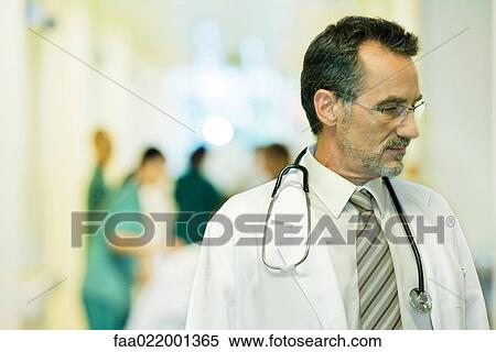 Stock Photography - Male doctor looking away, head and shoulders, hospital corridor in background. Fotosearch