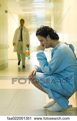 Stock Image - Male patient crouching in hospital corridor, holding head, looking down, doctor walking in background. Fotosearch