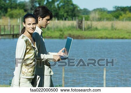 Stock Image - Man and woman standing by lake, man holding laptop, woman looking at camera. Fotosearch