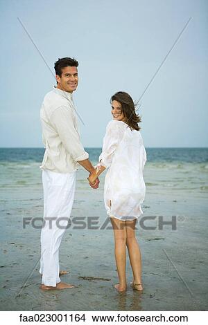 Man and young female companion on beach, holding hands, looking over shoulders at camera, full length View Large Photo Image Picture - Man and young female companion on beach, holding hands, looking over shoulders at camera, full length. Fotosearch