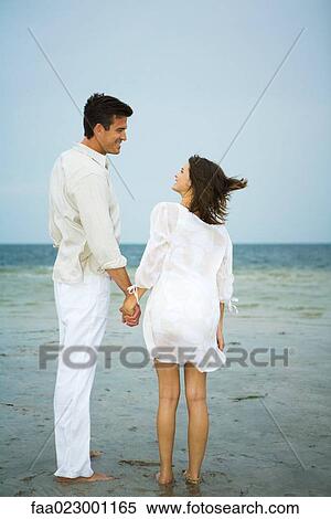 Man and young female companion on beach, holding hands, looking at each other, full length View Large Photo Image Stock Photography - Man and young female companion on beach, holding hands, looking at each other, full length. Fotosearch