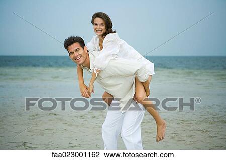 Man carrying young female companion piggyback on beach, smiling at camera View Large Photo Image Stock Image - Man carrying young female companion piggyback on beach, smiling at camera. Fotosearch