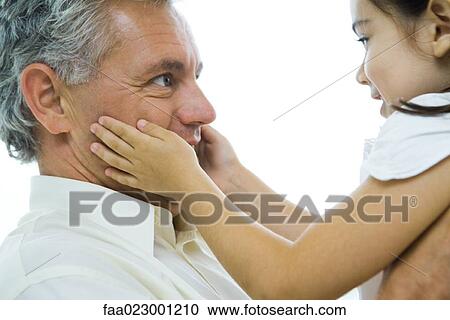 Stock Image - Man holding up little girl, girl's hands on man's cheeks, close-up. Fotosearch