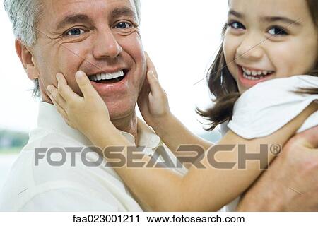 Stock Image - Man holding up little girl, girl's hands on man's cheeks, both smiling at camera, close-up. Fotosearch
