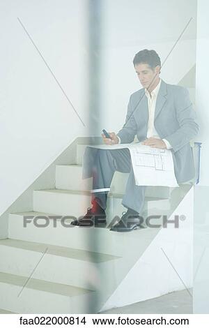 Picture - Man in suit sitting on stairs, looking at blueprints, using cell phone, seen through glass. Fotosearch