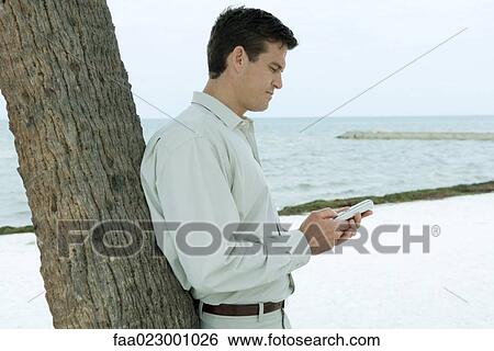 Stock Photograph - Man leaning against tree trunk at the beach, looking at cell phone. Fotosearch