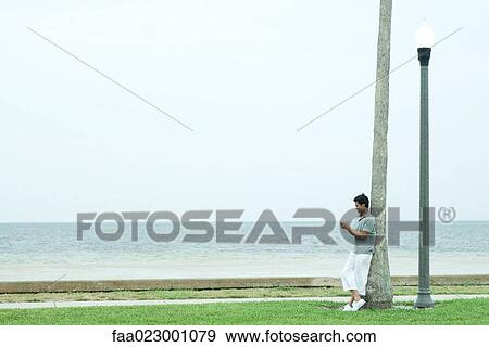 Man leaning against tree trunk, looking down at cell phone, full length View Large Photo Image Stock Photo - Man leaning against tree trunk, looking down at cell phone, full length. Fotosearch