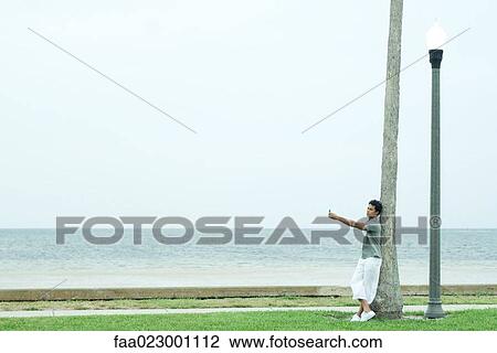 Man leaning against tree trunk, near beach, photographing self with cell phone View Large Photo Image Stock Image - Man leaning against tree trunk, near beach, photographing self with cell phone. Fotosearch