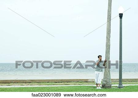 Man leaning against tree trunk, using cell phone, full length View Large Photo Image Stock Photo - Man leaning against tree trunk, using cell phone, full length. Fotosearch