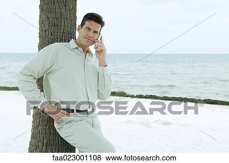 Man leaning against tree trunk, using cell phone, squinting at camera, hand on hip View Large Photo Image Stock Photo - Man leaning against tree trunk, using cell phone, squinting at camera, hand on hip. Fotosearch