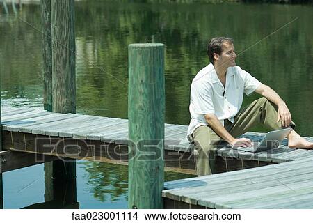 Man sitting on dock, using laptop computer, looking away View Large Photo Image Picture - Man sitting on dock, using laptop computer, looking away. Fotosearch
