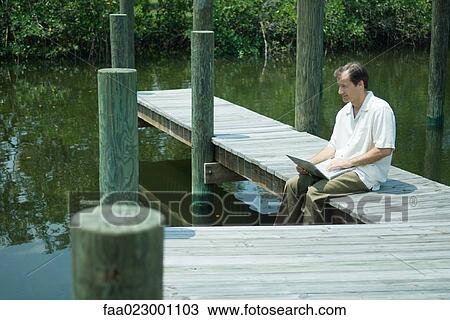 Man sitting on dock, using laptop computer, looking away, smiling View Large Photo Image Stock Image - Man sitting on dock, using laptop computer, looking away, smiling. Fotosearch