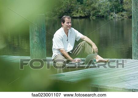 Man sitting on dock using laptop computer, looking away, view through foliage View Large Photo Image Stock Photo - Man sitting on dock using laptop computer, looking away, view through foliage. Fotosearch