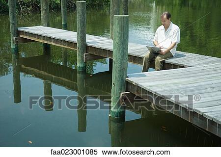 Man sitting on dock, using laptop computer, looking down, smiling View Large Photo Image Stock Photography - Man sitting on dock, using laptop computer, looking down, smiling. Fotosearch