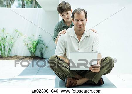 Man sitting on ground using laptop computer, son looking over his shoulder View Large Photo Image Stock Image - Man sitting on ground using laptop computer, son looking over his shoulder. Fotosearch