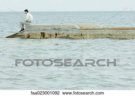 Stock Image - Man sitting on pier, using laptop computer, full length, side view. Fotosearch