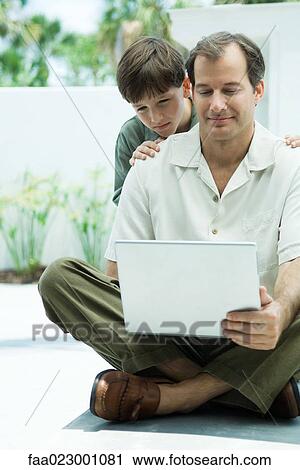 Stock Image - Man sitting on the ground using laptop computer, son looking over his shoulder. Fotosearch