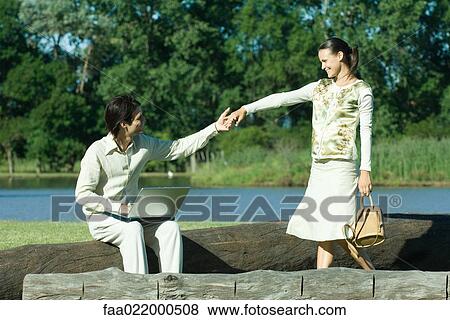 Man sitting on tree trunk with laptop on lap, reaching for woman's hand as she approaches View Large Photo Image Stock Photo - Man sitting on tree trunk with laptop on lap, reaching for woman's hand as she approaches. Fotosearch