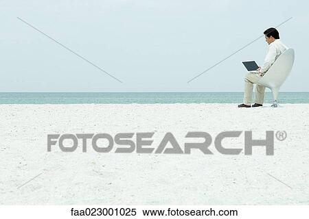 Stock Photography - Man using laptop computer at the beach, full length. Fotosearch