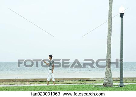 Stock Image - Man walking on sidewalk at the beach, looking at cell phone. Fotosearch