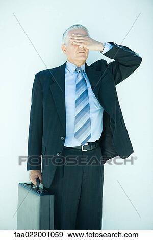 Stock Photo - Mature businessman holding briefcase, hand covering eyes, three quarter length. Fotosearch