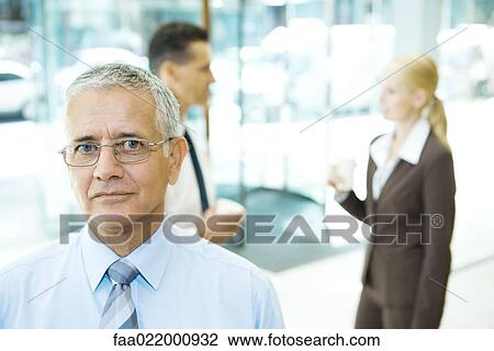 Stock Image - Mature businessman looking at camera, portrait, associates in background. Fotosearch