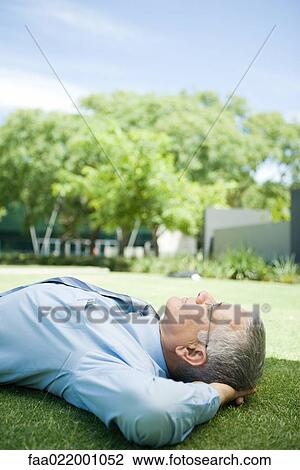 Stock Image - Mature businessman lying on ground outdoors, hands behind head, eyes closed. Fotosearch