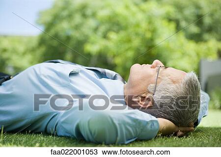 Stock Image - Mature businessman lying on ground outdoors, hands behind head, eyes closed, close-up. Fotosearch