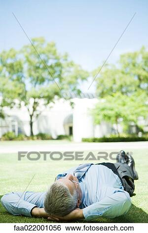 Stock Photograph - Mature businessman lying on ground outdoors, hands behind head, full length. Fotosearch