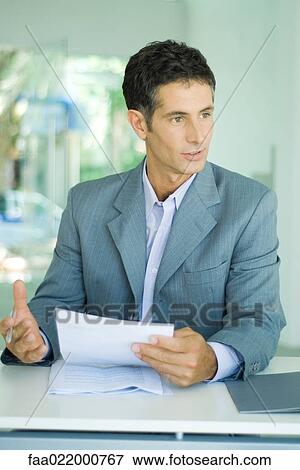 Mature businessman sitting at desk, holding document and speaking View Large Photo Image Stock Photo - Mature businessman sitting at desk, holding document and speaking. Fotosearch