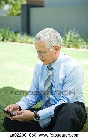 Stock Image - Mature businessman sitting on ground, looking at cell phone. Fotosearch
