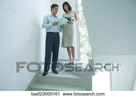 Stock Image - Mature businessman speaking to young female colleague on stairs, showing document. Fotosearch