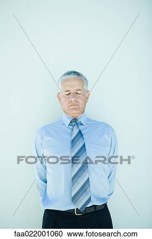Stock Image - Mature businessman standing, hands behind back, eyes closed. Fotosearch