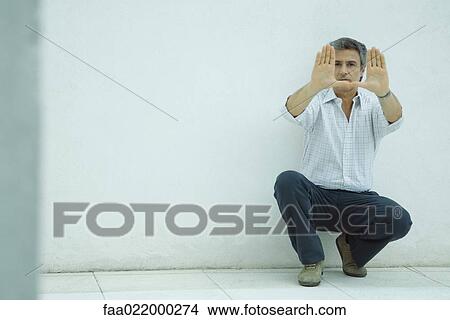 Mature man crouching, making frame with hands, looking at camera View Large Photo Image Picture - Mature man crouching, making frame with hands, looking at camera. Fotosearch