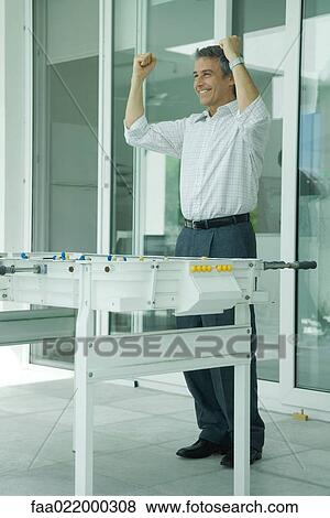 Mature man standing by foosball table, smiling and holding up fists, full length View Large Photo Image Stock Photo - Mature man standing by foosball table, smiling and holding up fists, full length. Fotosearch
