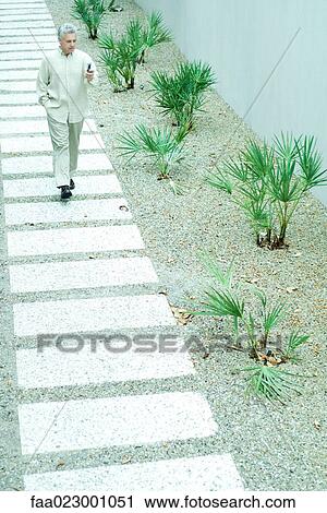 Stock Image - Mature man walking on path, looking at cell phone, full length. Fotosearch
