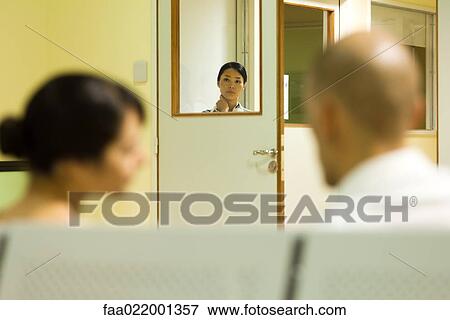 Stock Photo - Medical worker looking through window, adults sitting in hospital waiting room in foreground. Fotosearch