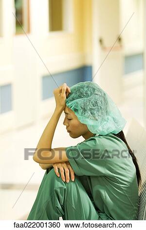 Stock Image - Medical worker sitting in hospital corridor, holding head, eyes closed. Fotosearch