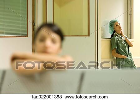 Stock Photo - Medical worker standing in hospital waiting room, hands in pockets, looking at camera, woman resting head on arms in foreground. Fotosearch