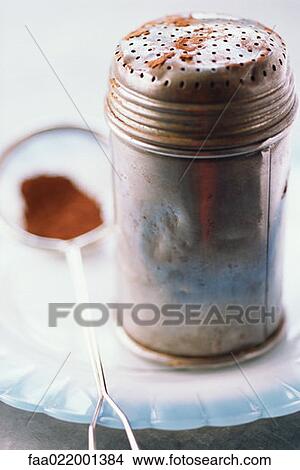Picture - Metal shaker containing cocoa and sieve on plate, close-up. Fotosearch