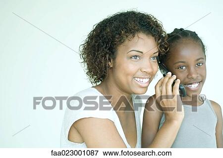 Stock Photo - Mother holding cell phone up to daughter's ear, both smiling, daughter looking at camera. Fotosearch