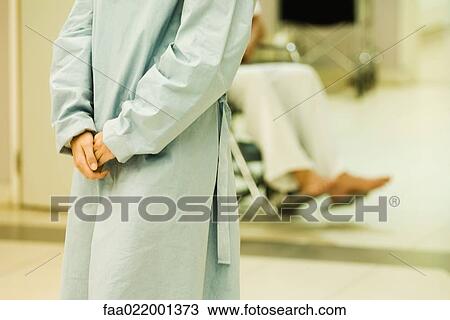 Stock Image - Patient standing with clasped hands, wearing hospital gown, cropped view. Fotosearch