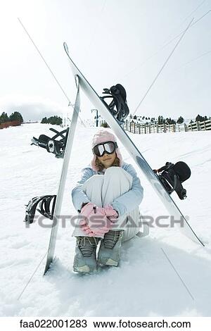 Stock Image - Preteen girl sitting on snow underneath propped up snowboards. Fotosearch
