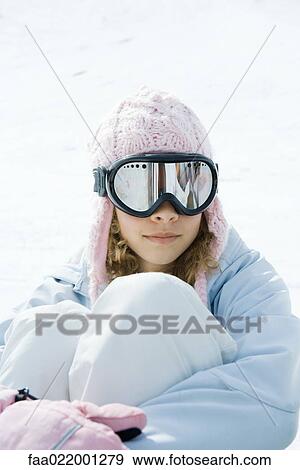 Stock Photo - Preteen girl wearing ski gear, sitting in snow, hugging knees, portrait. Fotosearch
