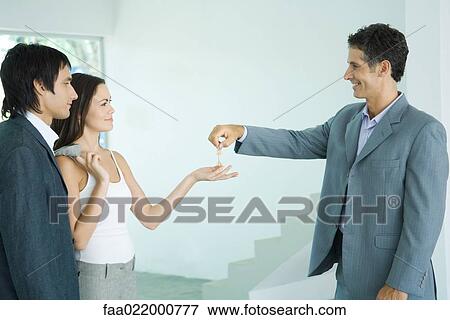 Real estate agent holding out keys to young couple, standing in bare home interior View Large Photo Image Stock Photo - Real estate agent holding out keys to young couple, standing in bare home interior. Fotosearch