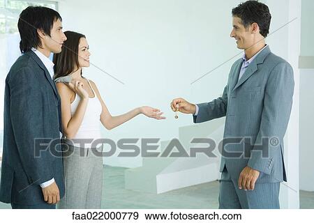 Real estate agent holding out keys to young couple, standing in bare home interior View Large Photo Image Stock Photo - Real estate agent holding out keys to young couple, standing in bare home interior. Fotosearch