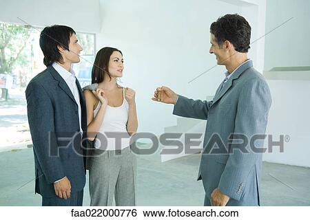 Real estate agent holding out keys to young couple, standing in bare home interior View Large Photo Image Stock Photograph - Real estate agent holding out keys to young couple, standing in bare home interior. Fotosearch