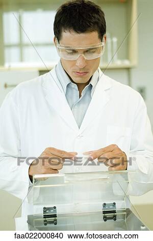 Stock Image - Researcher wearing protective goggles picking up Petri dish. Fotosearch