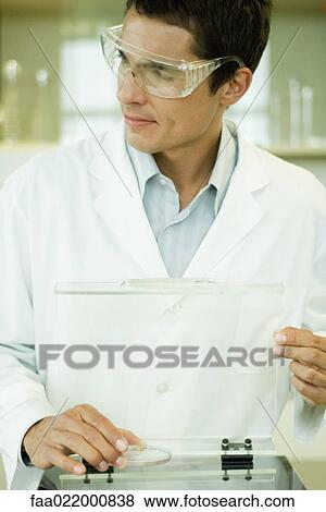 Stock Photo - Researcher wearing protective goggles picking up Petri dish, looking away. Fotosearch